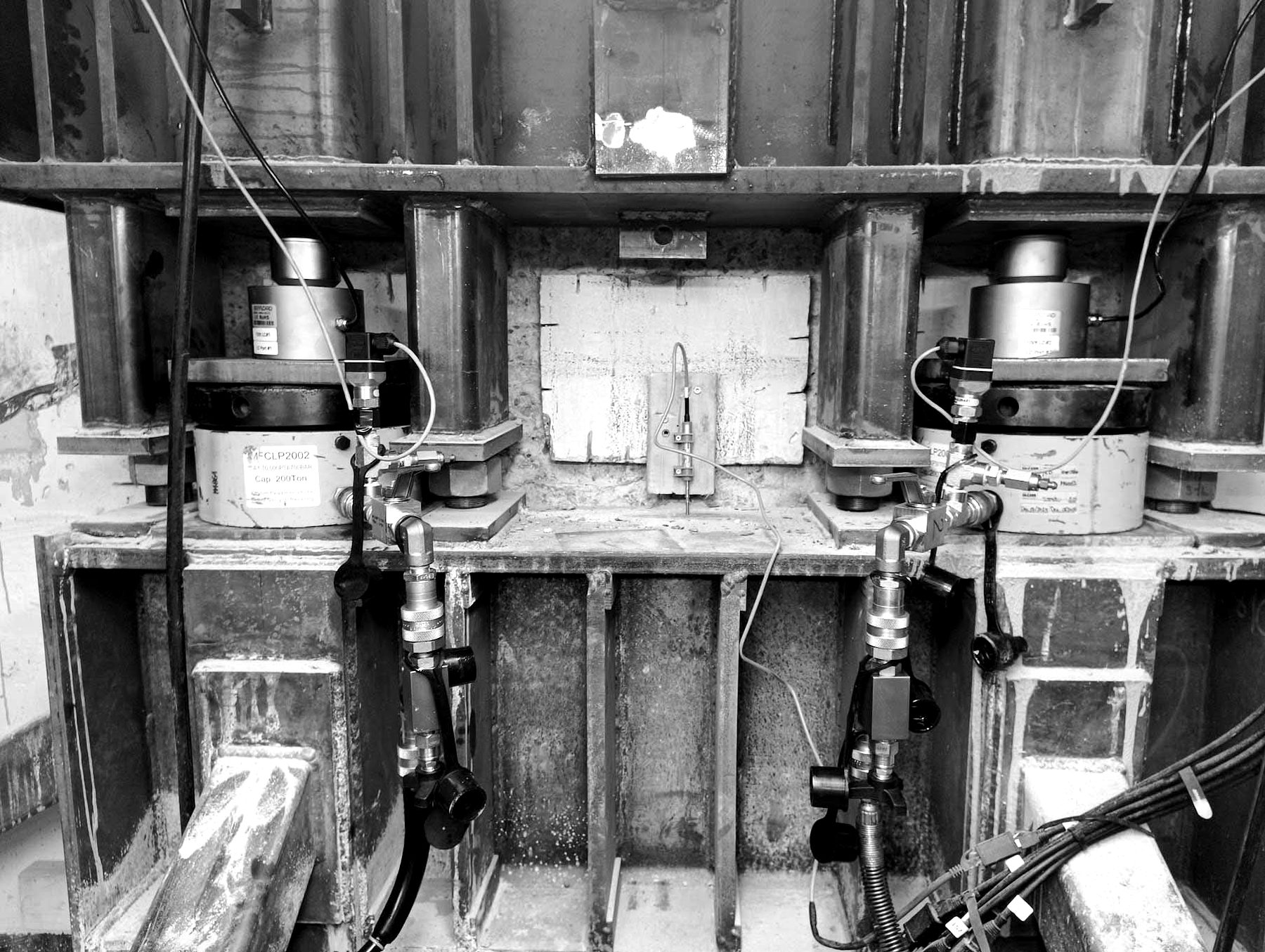 Two hydraulic cylinders with load cells on top, sitting on the stiffened lower bracket, with a displacement sensor on the column face between them